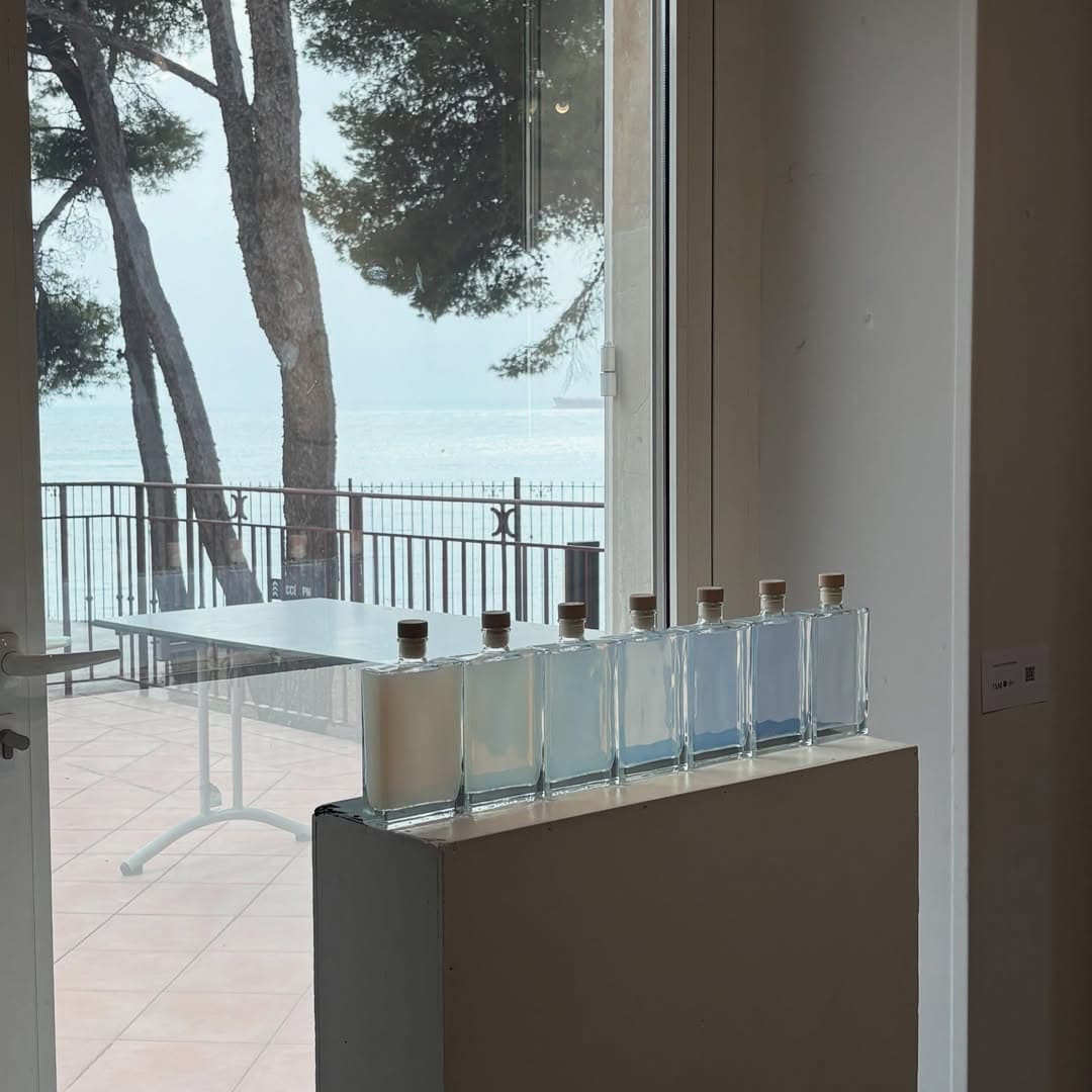 Seven glass bottles on a plinth by a window to the sea